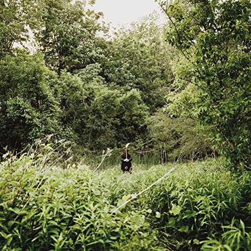 Person in black standing in tall green grass and trees.
