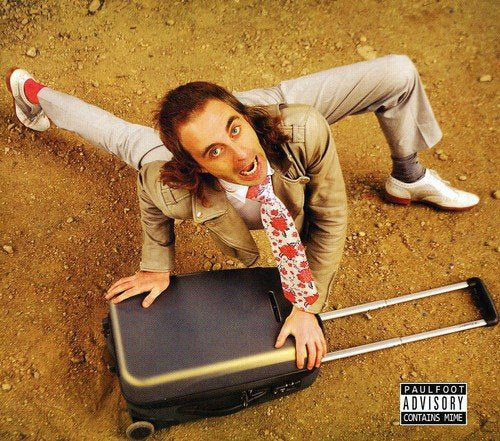 Man in suit contorting body around a black suitcase on dirt.