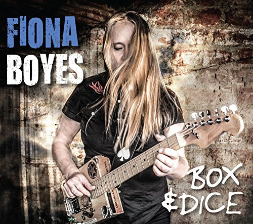 Fiona Boyes playing a blue electric guitar against a brick wall.