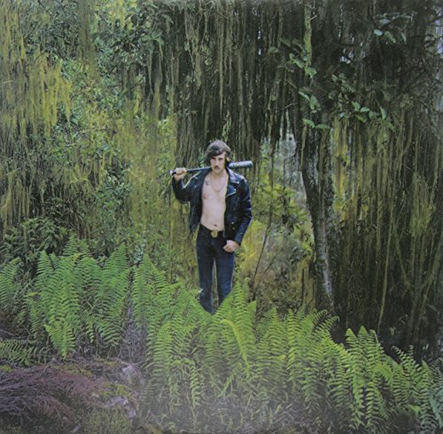 Man with a baseball bat stands in lush, overgrown greenery.
