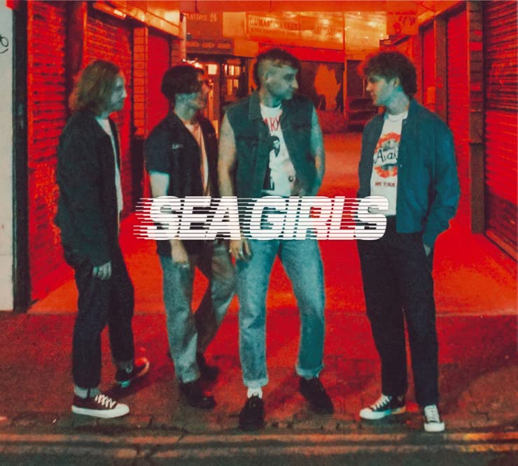 Four young men standing on a sidewalk at night with red lighting and Sea Girls text.
