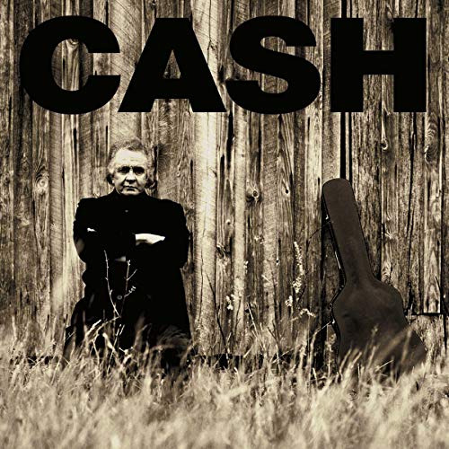 Johnny Cash sitting in tall grass before a wooden barn with a guitar case.
