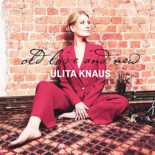 Woman in a red suit sitting on a patterned rug against a brick wall.