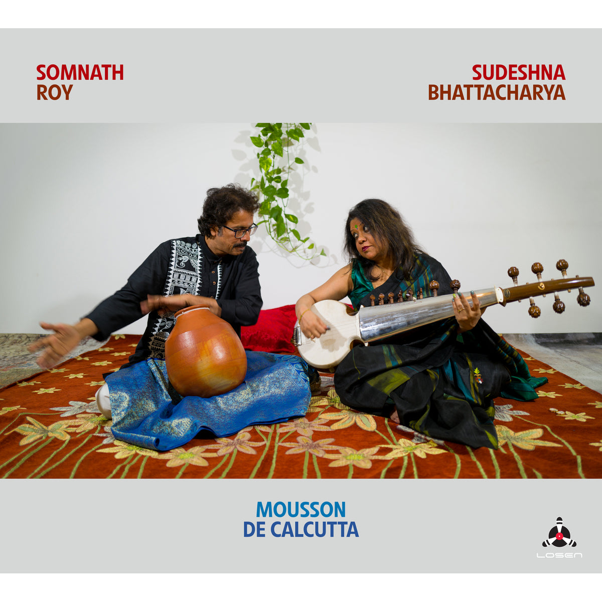 Two musicians with Indian instruments seated on a patterned rug.
