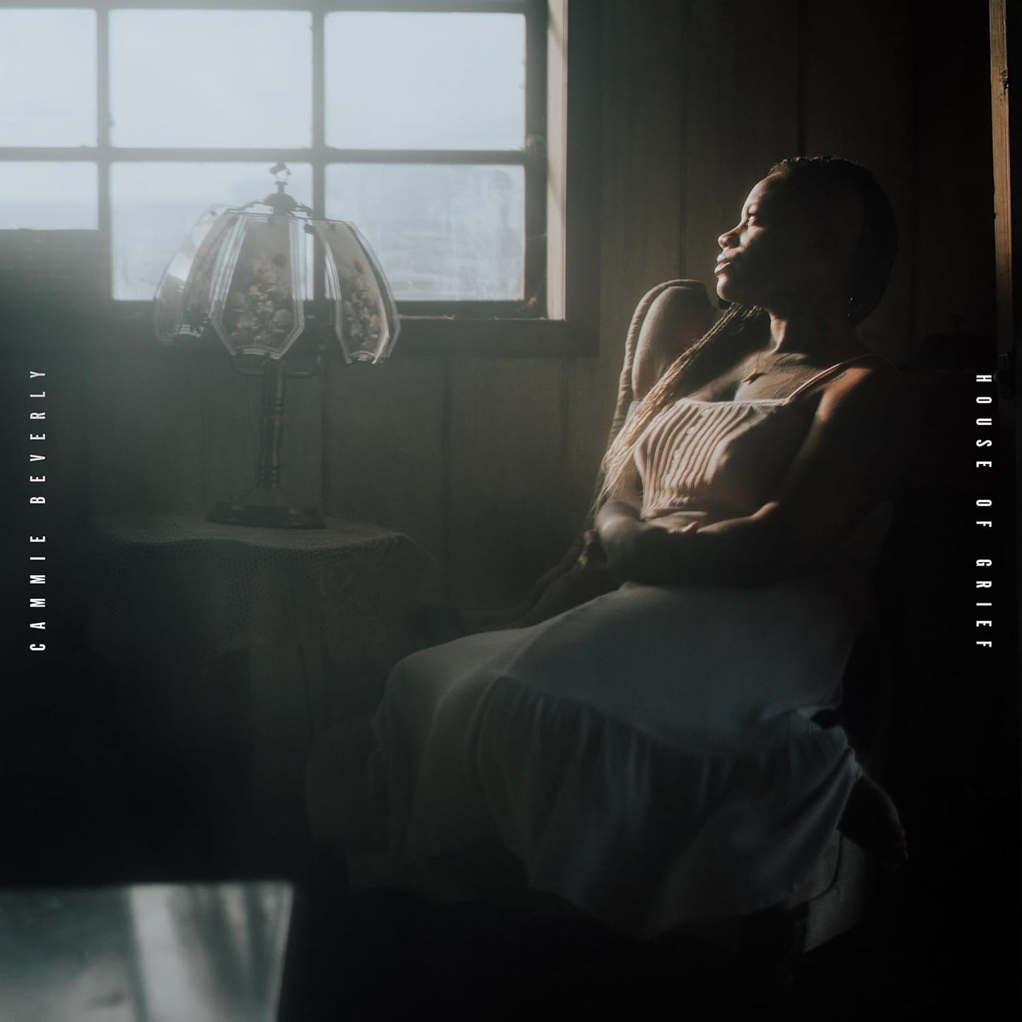 Woman seated near a window with a lit lamp, album cover.