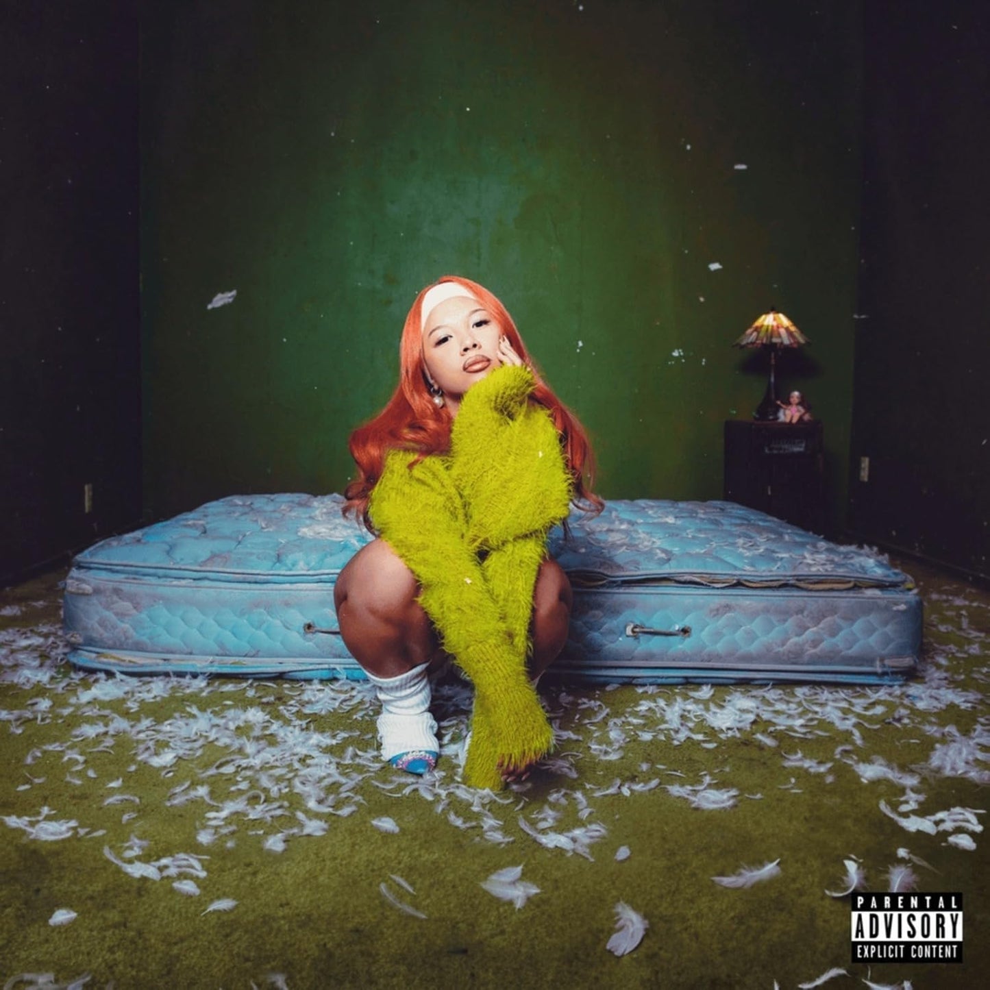 Woman with orange hair sitting on a mattress in a room.
