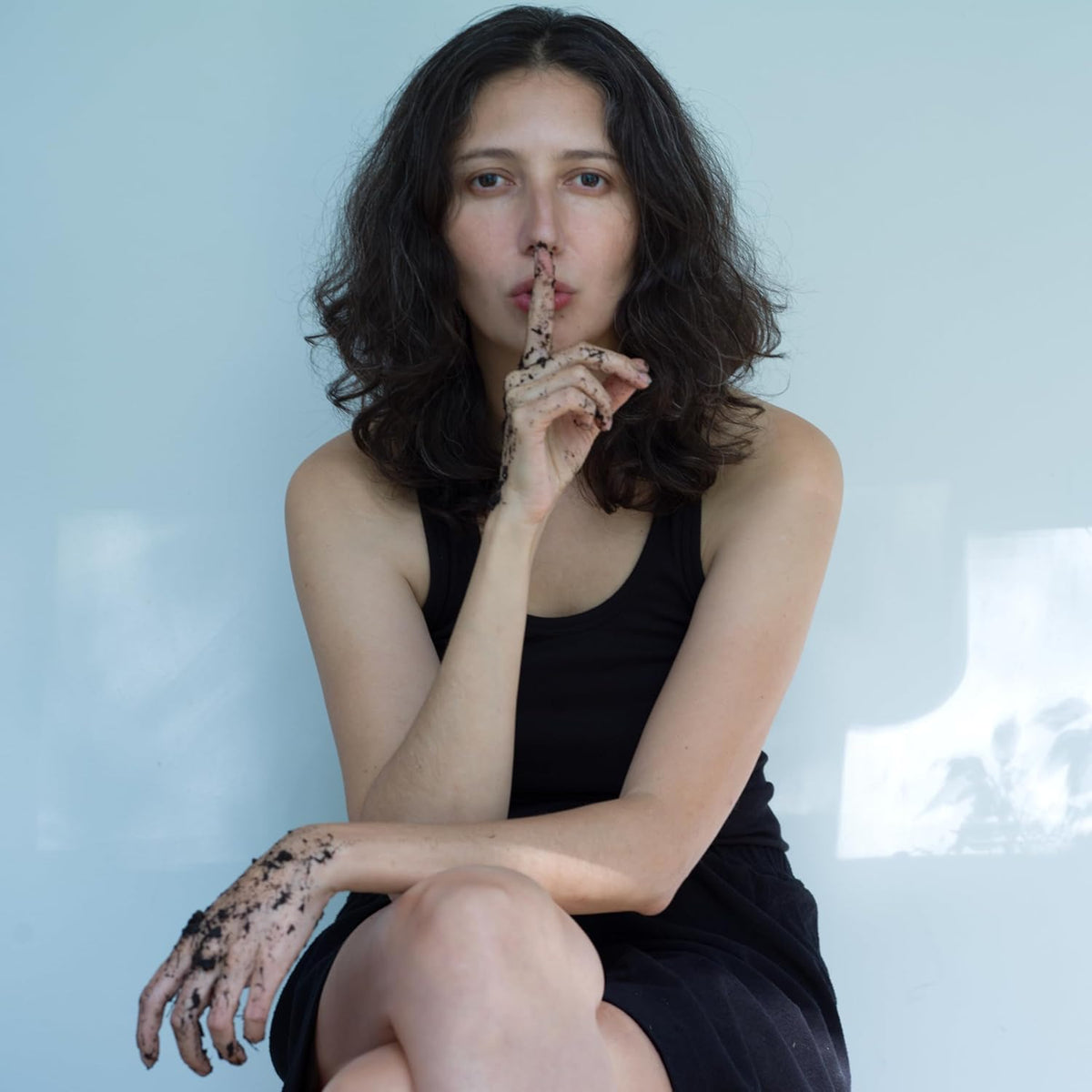Woman with dark soil on hands and arms, holding a finger to lips.