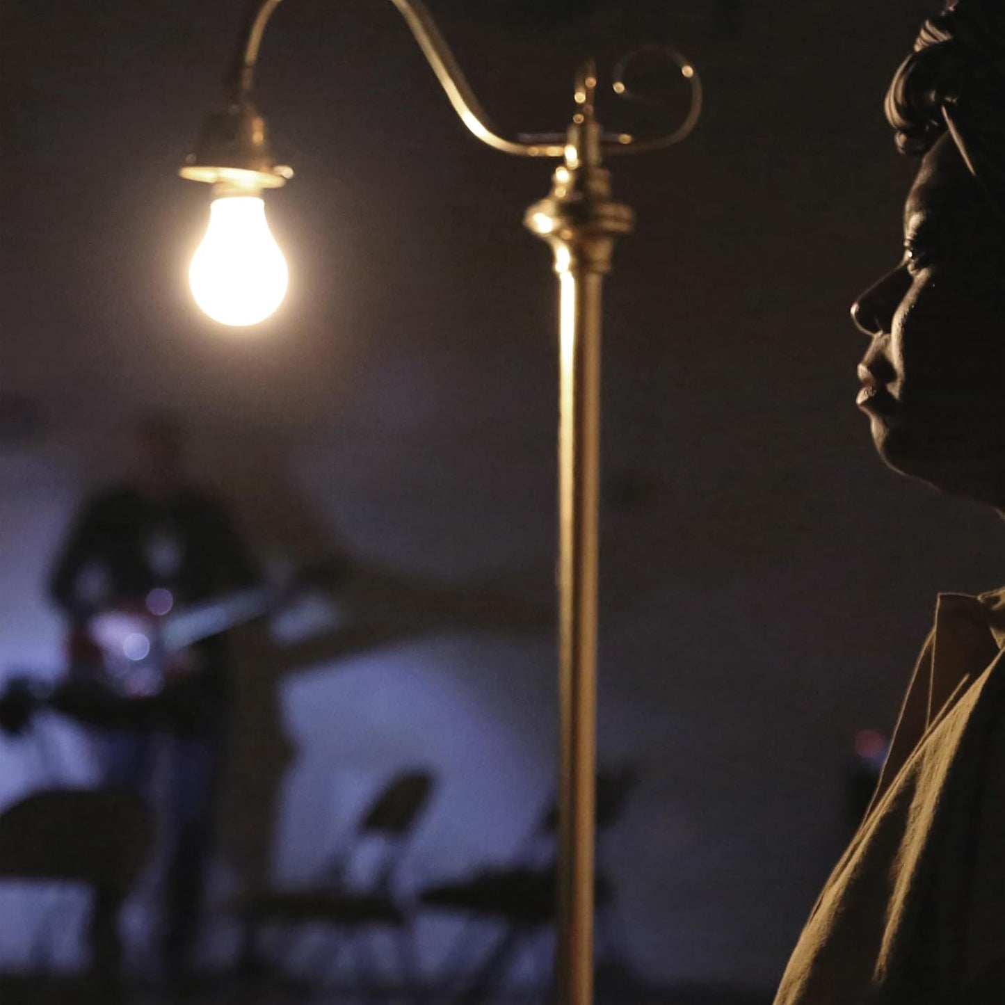 Man in profile beside a lamp, musicians blurred in background.