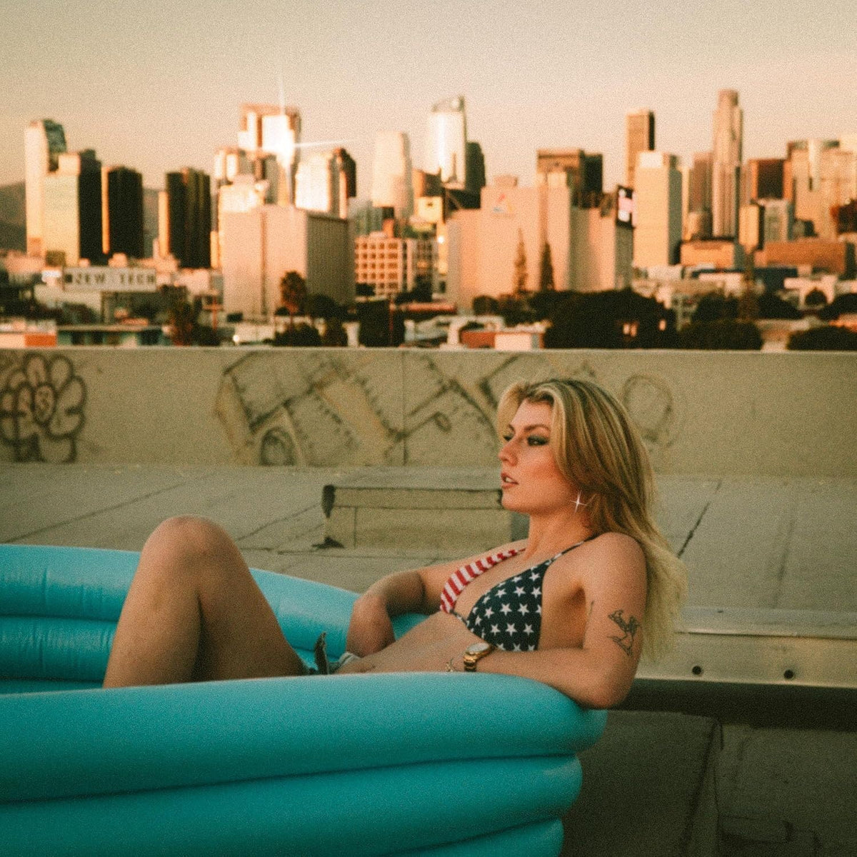 Blonde woman in inflatable pool on rooftop with city skyline.