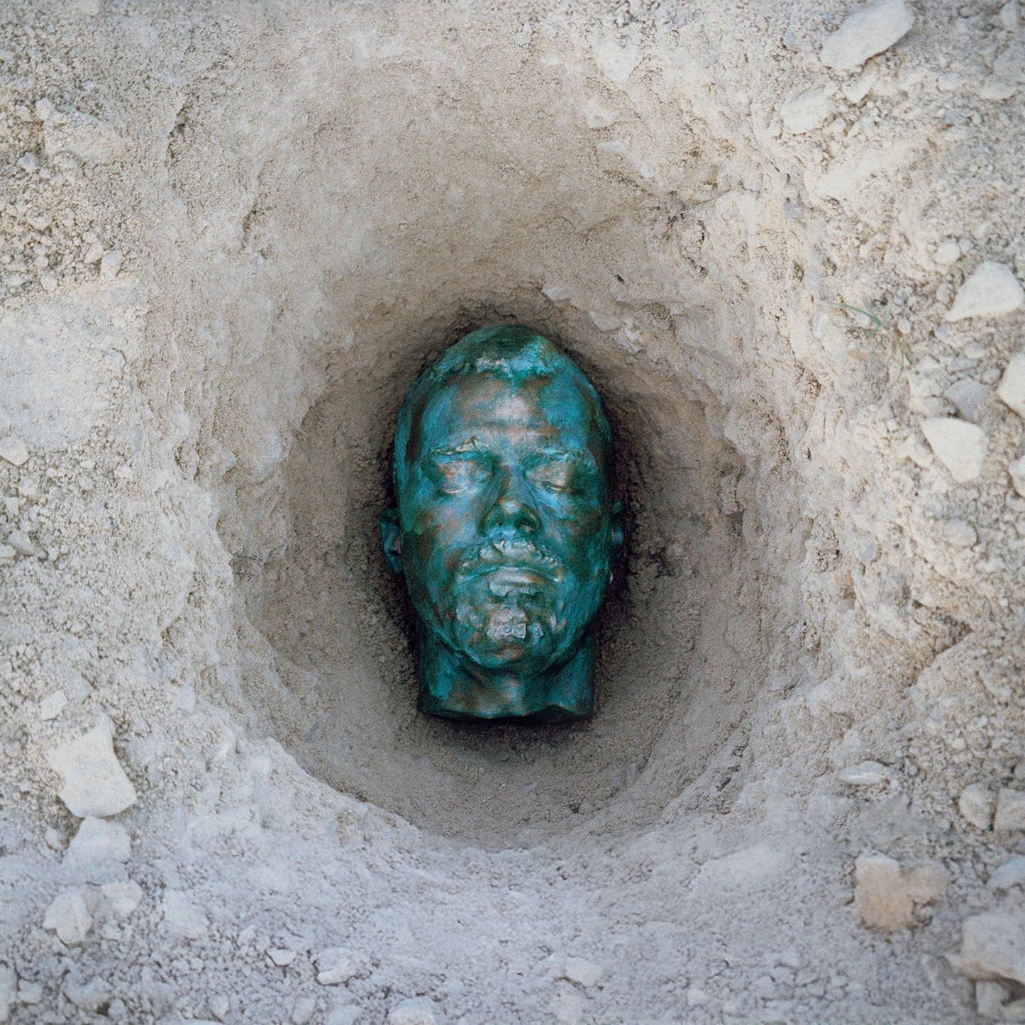 Bronze-colored human head sculpture partially buried in sandy ground.
