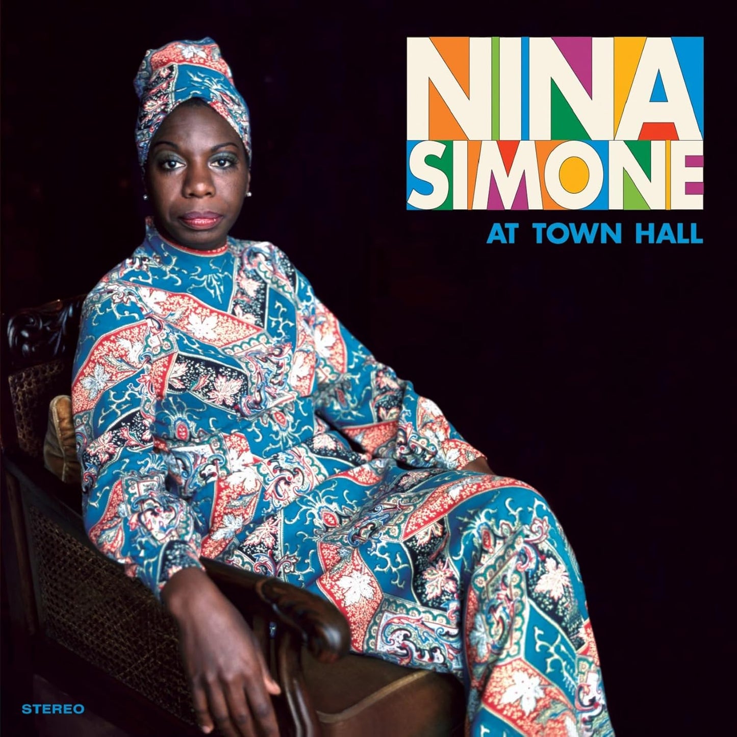 Nina Simone wearing a patterned headwrap and dress, seated in a chair against black.