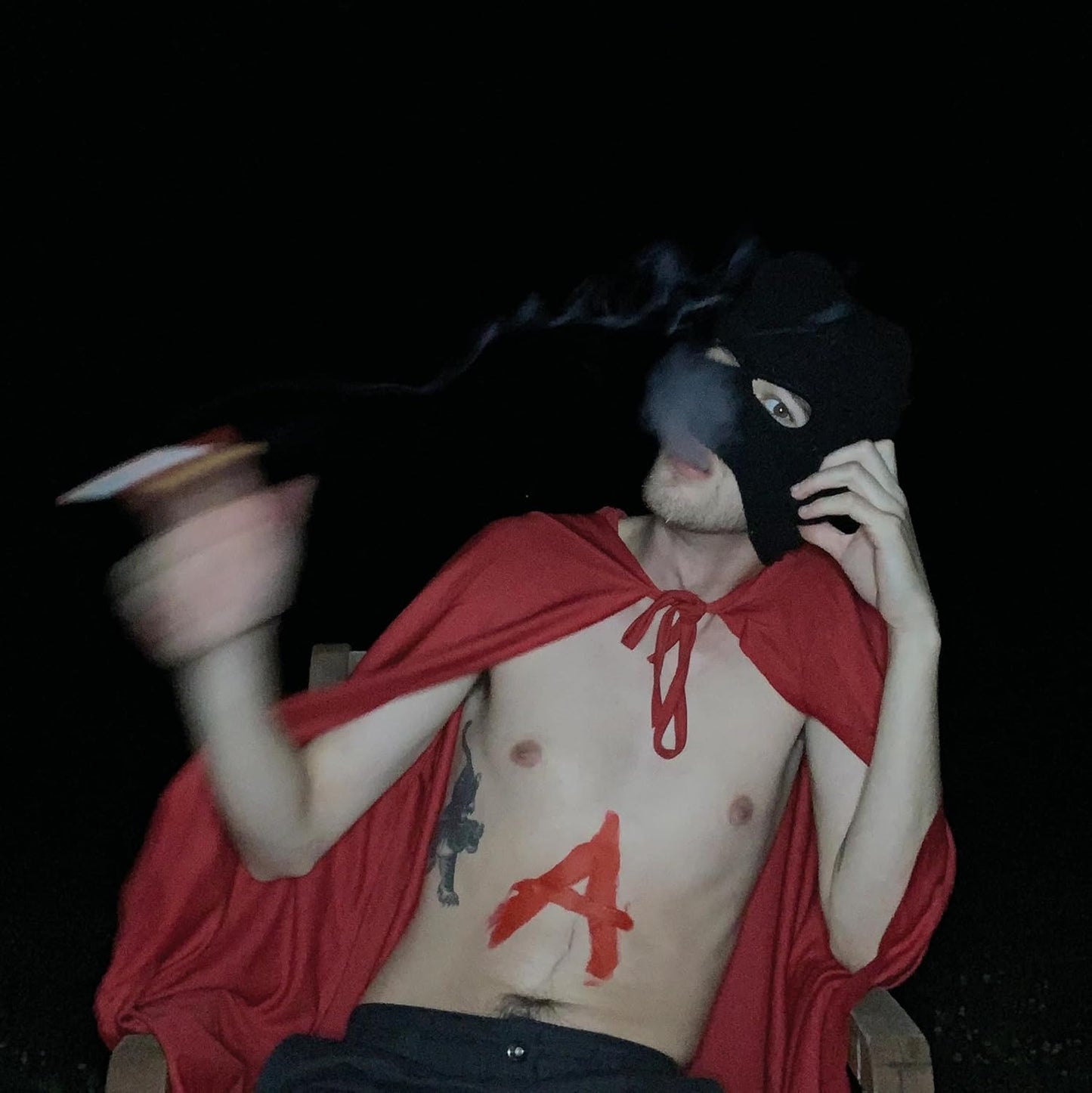 Person in a balaclava and red cape with a painted "A" on chest.