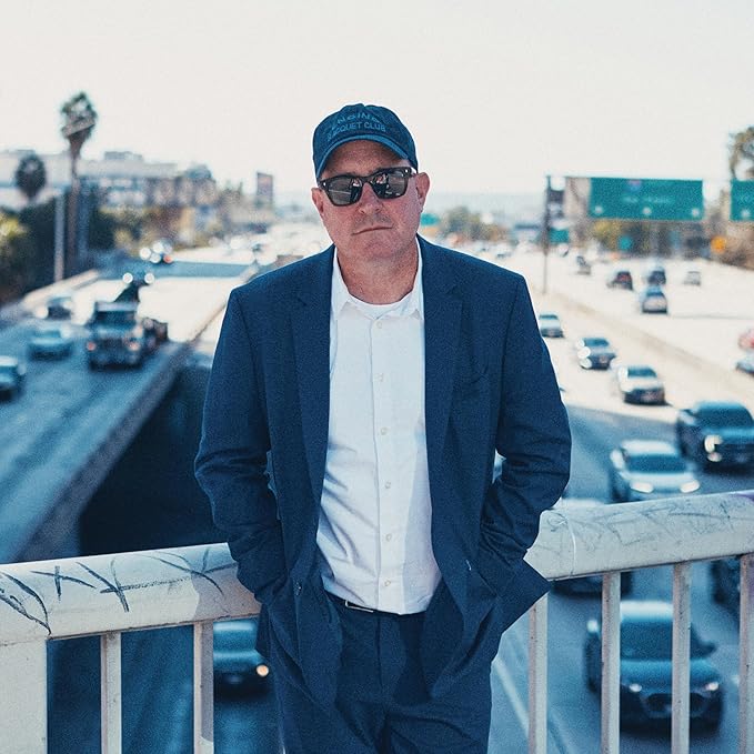 Craig Finn in suit and cap, standing on a bridge over traffic.