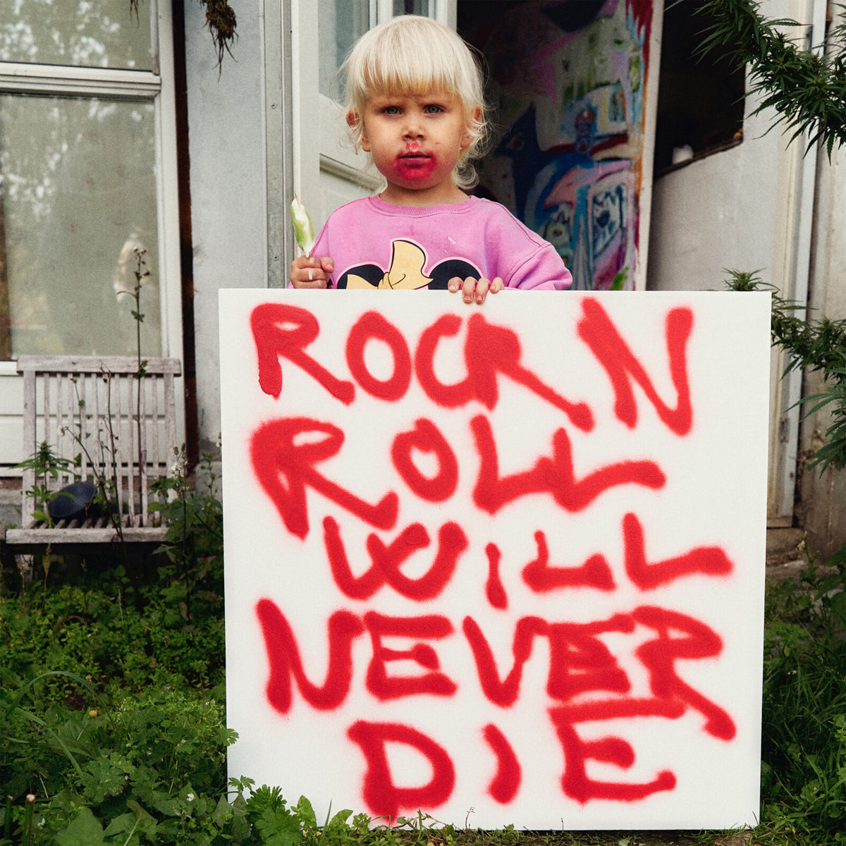 Child holding a sign with red graffiti text: “Rock N Roll Will Never Die.”