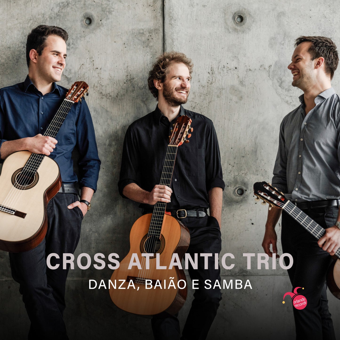 Three musicians with guitars stand against a textured gray wall.