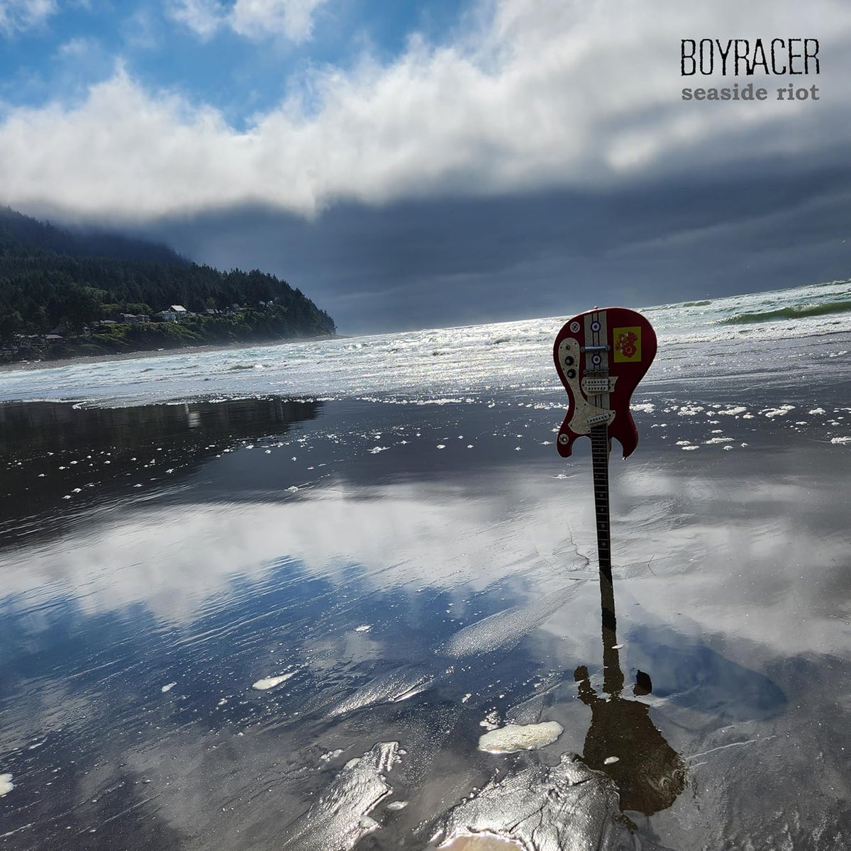 Red electric guitar standing on a wet, sandy beach with ocean backdrop.