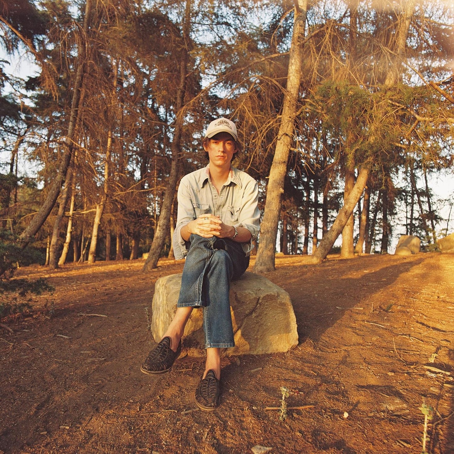Man in denim seated on a rock among trees.