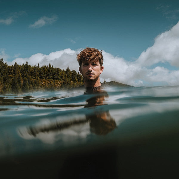 Man’s face partially submerged in water with trees and sky backdrop.
