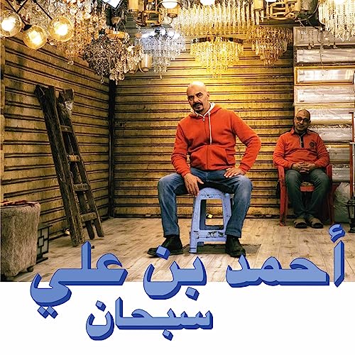 Two men seated indoors with chandeliers and Arabic text overlay.