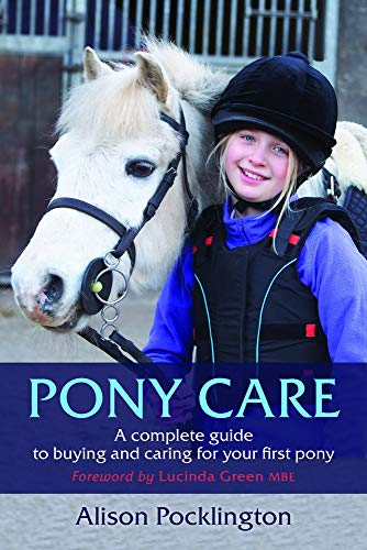 A smiling girl in riding gear stands next to a white pony.