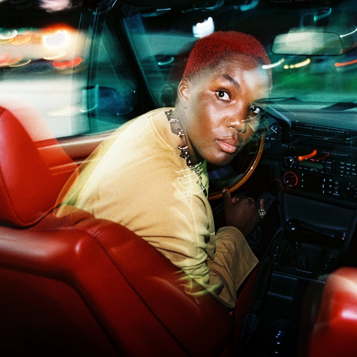 Arlo Parks with reddish hair, looking at the camera from a car.