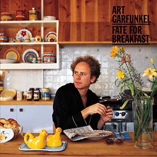 Art Garfunkel sits at a kitchen table with a newspaper and flowers.