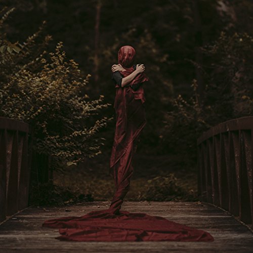 A person draped in red fabric stands on a wooden bridge in a forest.