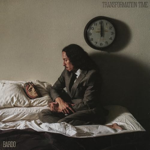 Person in suit sits on bed with mask and clock.