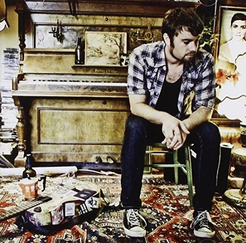 Man with guitar sits before an antique piano in a room.