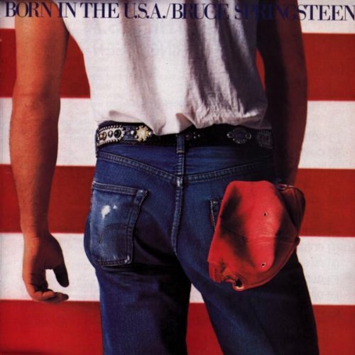 A man in a white t-shirt and jeans standing before an American flag background.