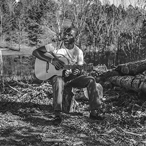 Black and white photo of Cedric Burnside sitting on a stump playing acoustic guitar.