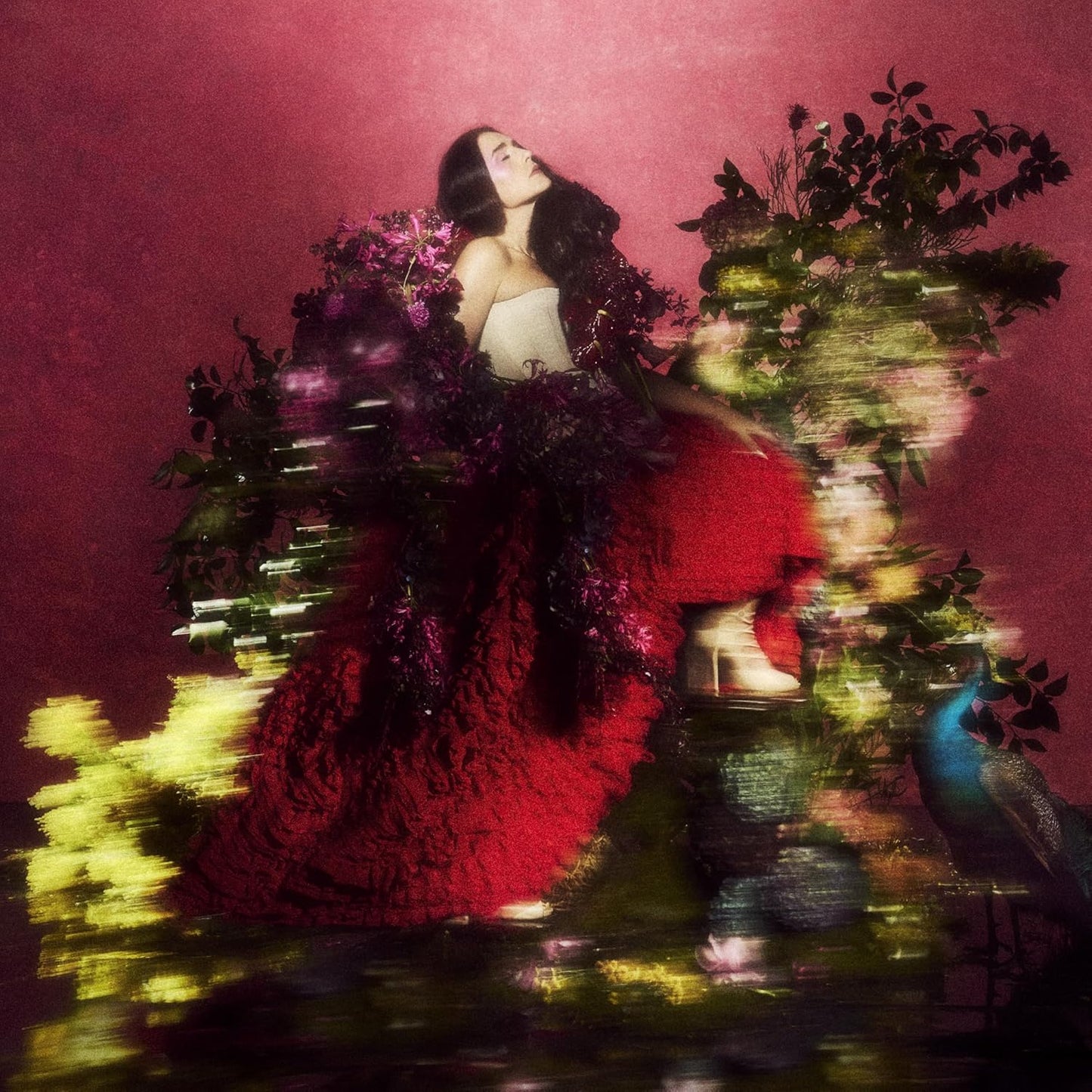 Jessie Ware in a voluminous red dress surrounded by dark flowers against pink background.