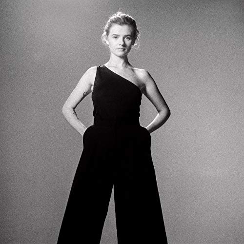 The image shows a woman in a black jumpsuit standing with her hands on her hips. For accessibility purposes, a suitable alt text could be: "Woman in a black one-shoulder jumpsuit posing against a grey background."