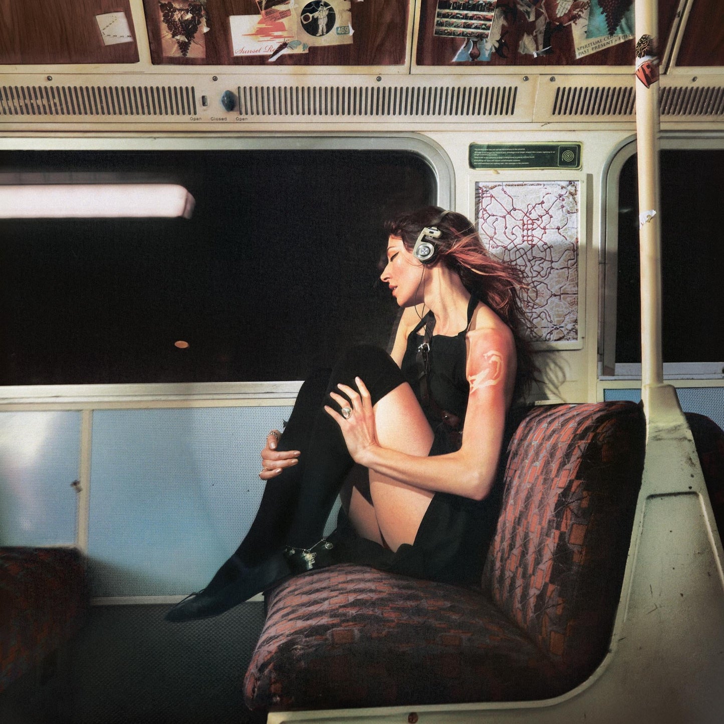 Caroline Polachek sitting on a train seat wearing headphones and a black dress.