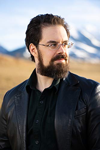 A bearded man with glasses wearing a black jacket against a mountain landscape background.