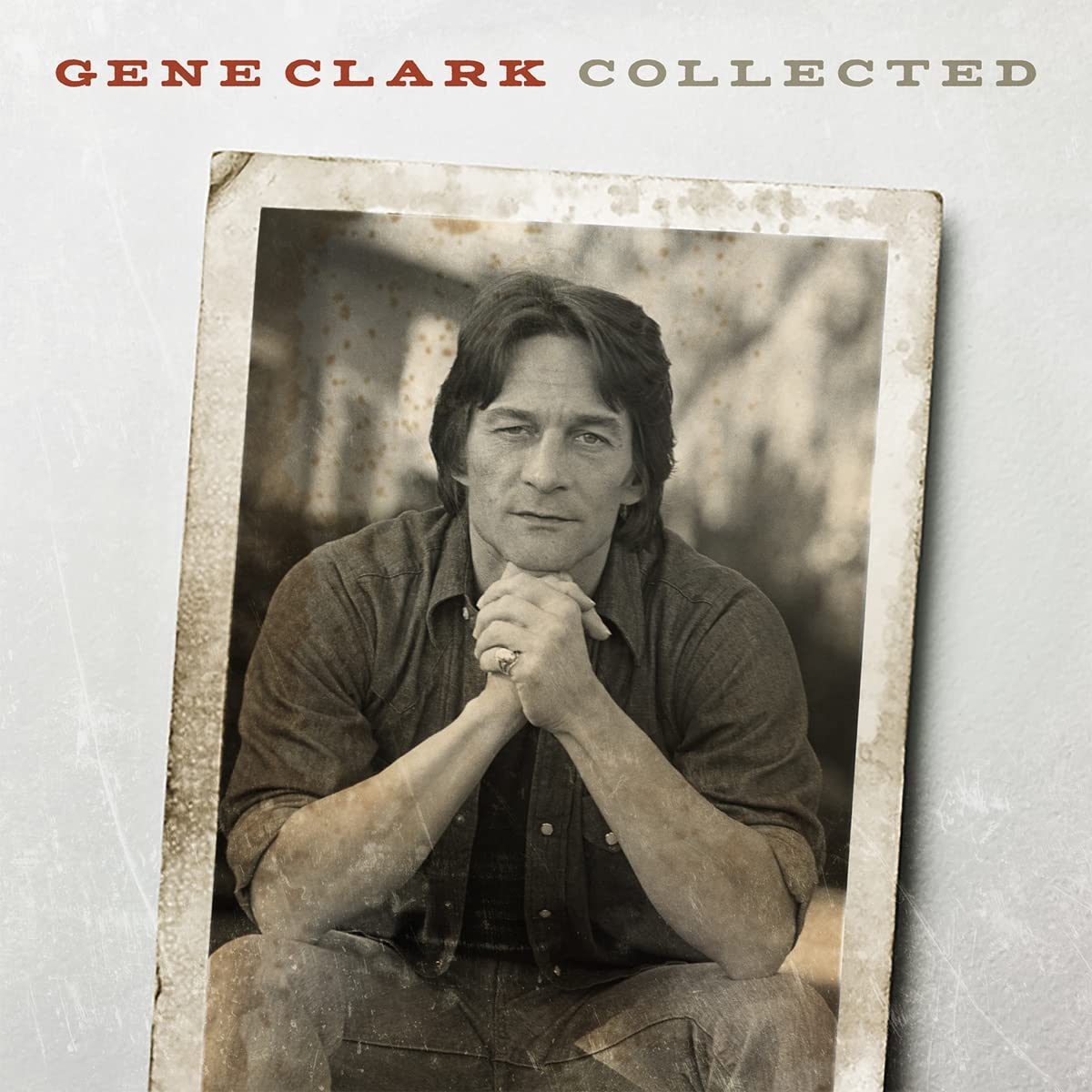Gene Clark sitting with hands clasped, wearing a dark shirt and jeans, sepia photo.