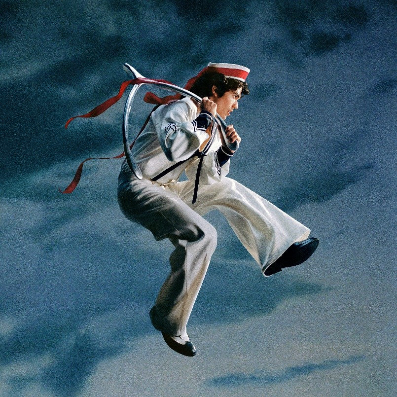 Conan Gray in a sailor suit mid-air against a cloudy blue background.