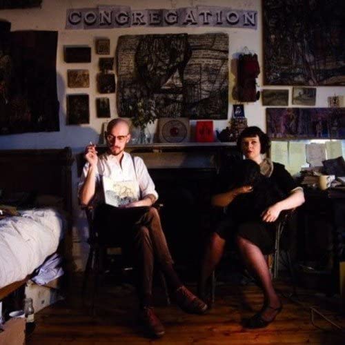 Album cover featuring two people and artwork in a cluttered room.