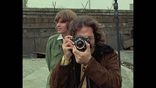 Two men with a vintage camera stand before a brick wall.
