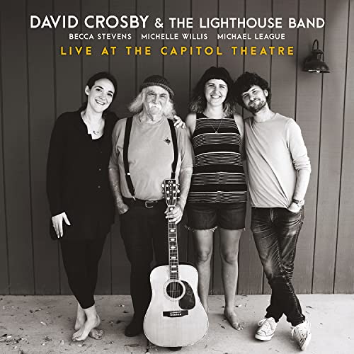 Four musicians standing together with a guitar, wearing casual clothing, on concrete.