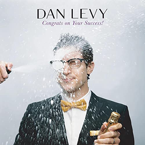 Dan Levy wearing a tuxedo and bowtie, being sprayed with champagne against a grey background.