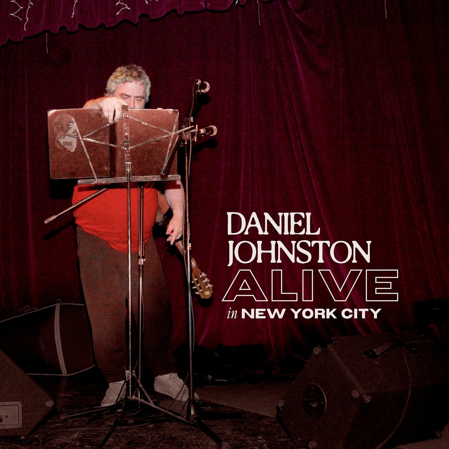 Daniel Johnston performing on stage in front of a dark red curtain.