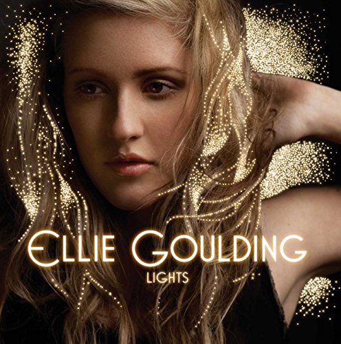 Ellie Goulding portrait surrounded by glowing golden light particles against a black background.