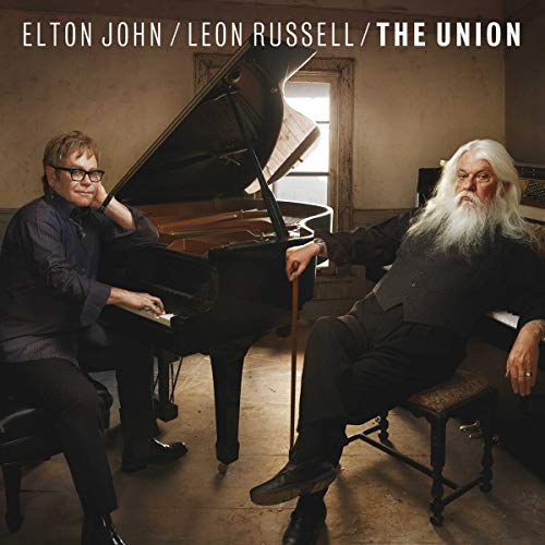 Elton John and Leon Russell sitting by a grand piano in a rustic room.