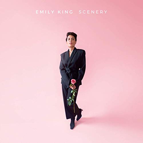 Emily King stands against a pink background wearing a black suit holding a rose.