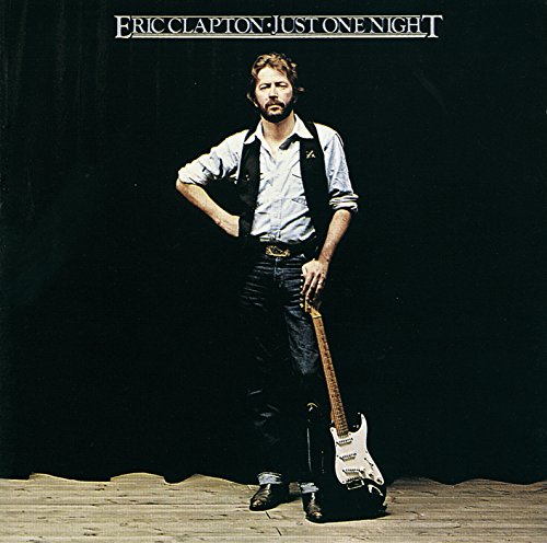 Eric Clapton standing on a stage holding a guitar against a black curtain background.