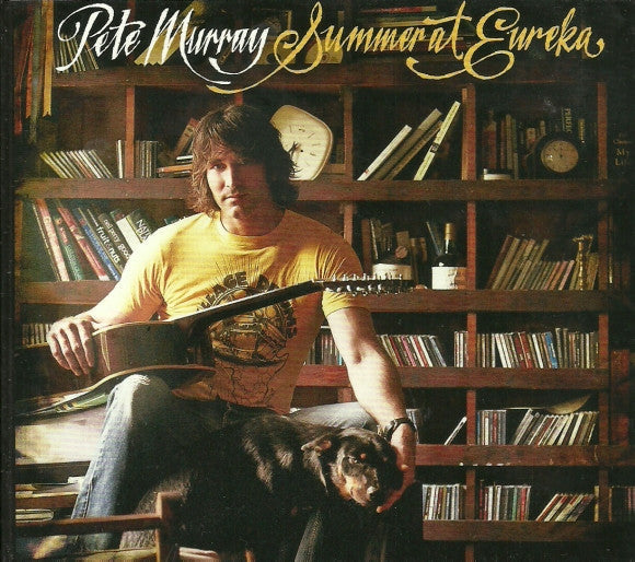 Man with guitar and dog seated before a full bookshelf.