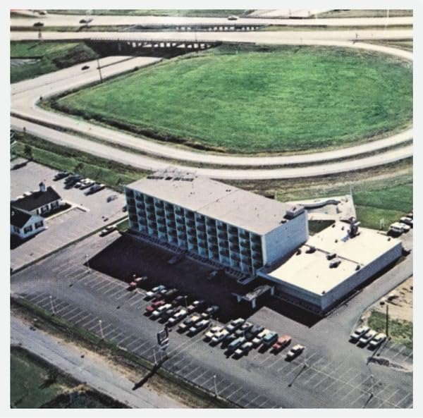 Aerial view of a low-rise hotel and parking lot with surrounding greenery.
