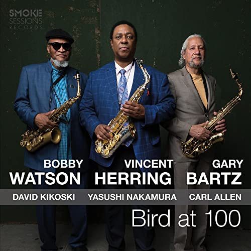 Three jazz musicians holding saxophones stand against a dark green wall.