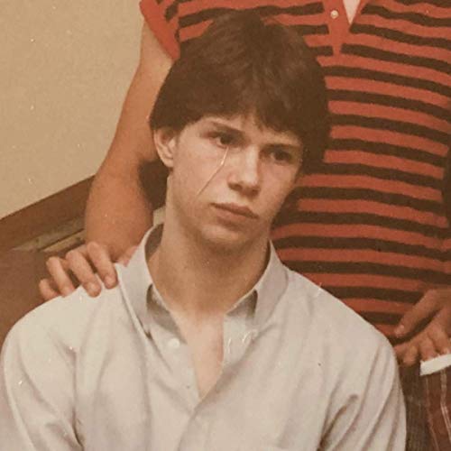 A young man with dark hair in a light shirt, with a hand on his shoulder.
